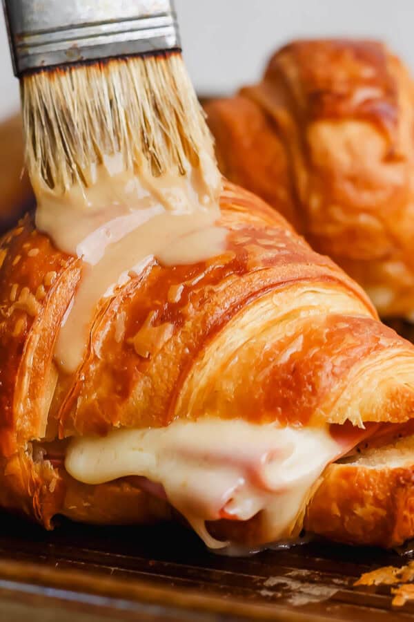 A melted ham and cheese croissant on a baking sheet with a brush adding some honey mustard sauce on top.