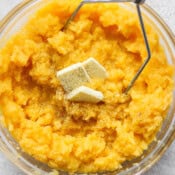 A large glass bowl filled with mashed rutabaga with butter on top and a potato masher sticking out.
