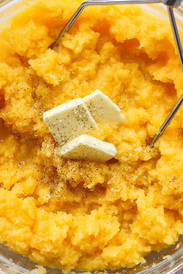 A large glass bowl filled with mashed rutabaga with butter on top and a potato masher sticking out.
