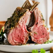 Two racks of lamb on a plate leaning on each other with herbed oil on top.