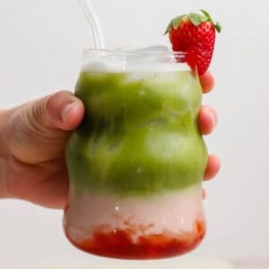 Someone holding a glass of strawberry match latte with a strawberry garnish and glass straw sticking out.
