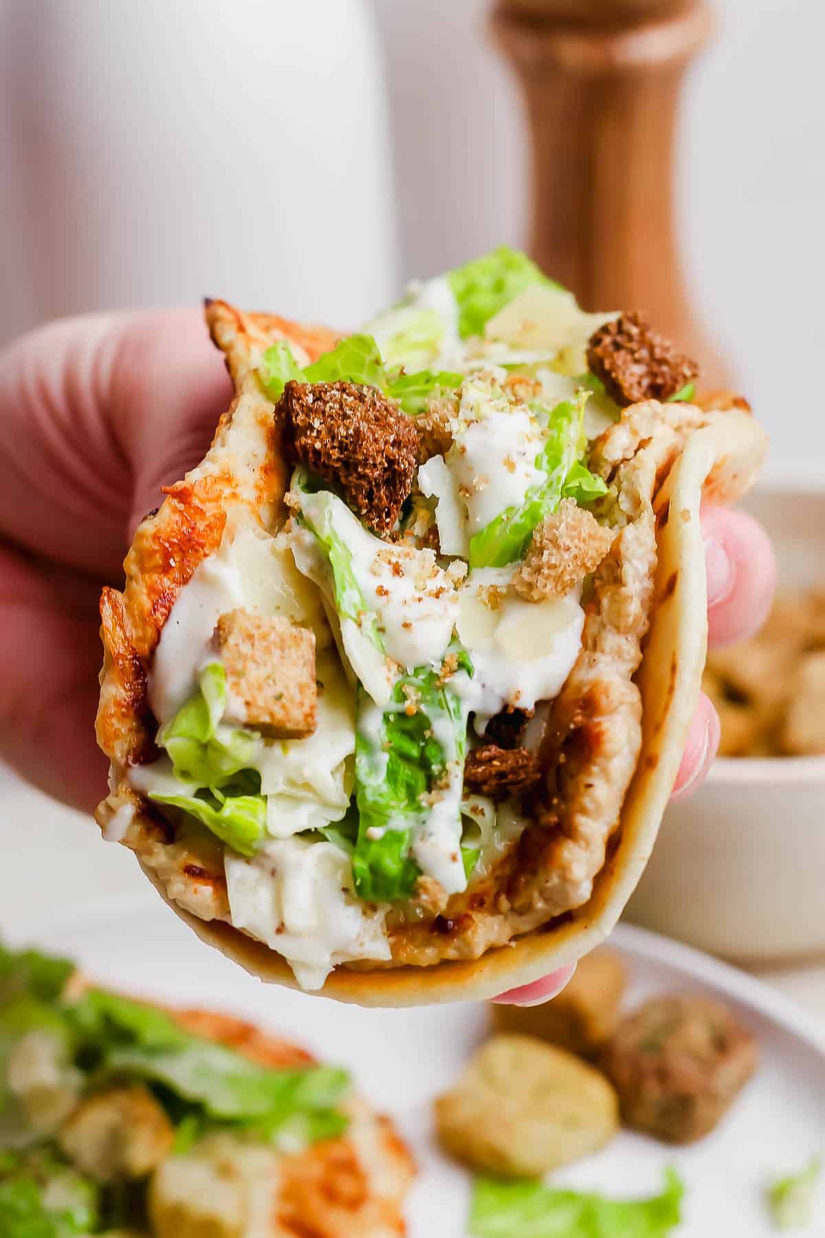 A hand holding a Chicken smash burger taco topped with croutons.