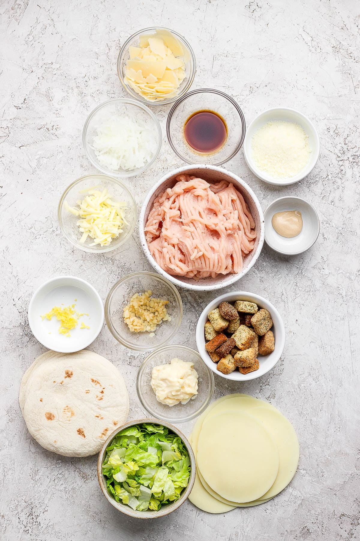Individual bowls of shaved parmesan cheese, shaved onion, grated parmesan cheese, grated butter, ground chicken, dijon mustard, minced garlic, lemon zest, croutons, mayo, chopped romaine, tortillas, white cheese, and worcestershire.