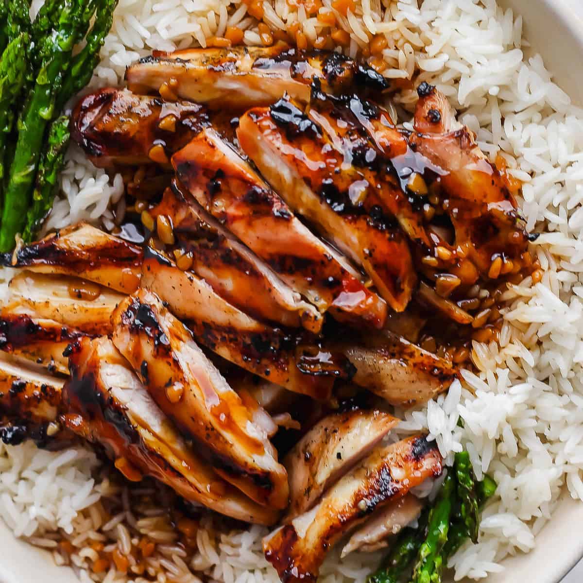 A close up of a plate of rice with grilled asparagus and grilled bourbon chicken on top.
