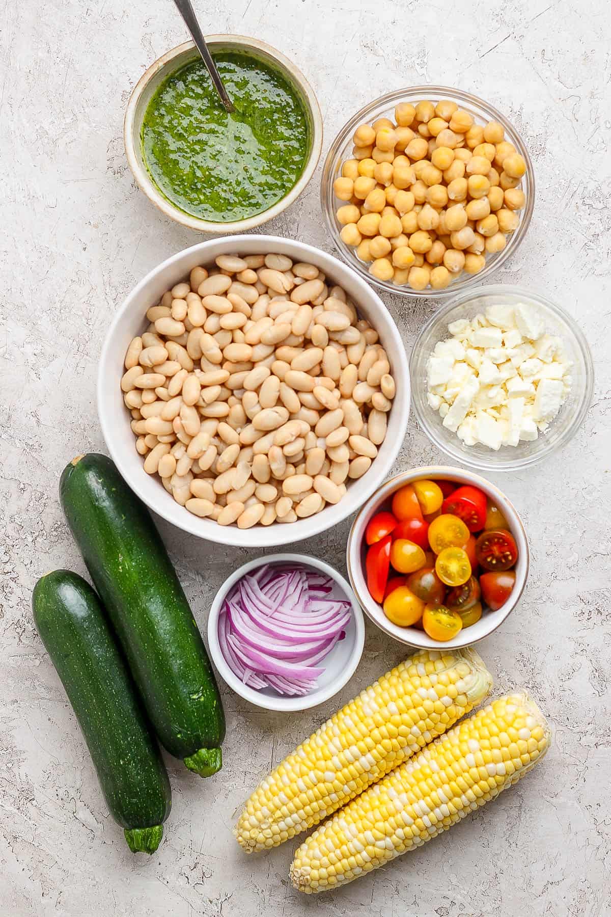 Individual bowls of chimichurri sauce, garbonzo beans, canellini beans, feta cheese, cherry tomatoes, red onion, zuchinni, and corn on the cob.