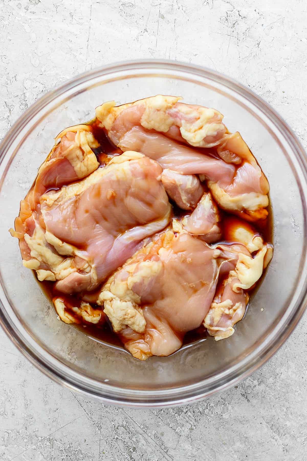 Chicken thighs marinating in a large glass bowl with soy sauce.