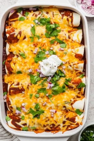 A top shot of casserole dish filled with pulled pork enchiladas topped with cilantro, red onion and sour cream.