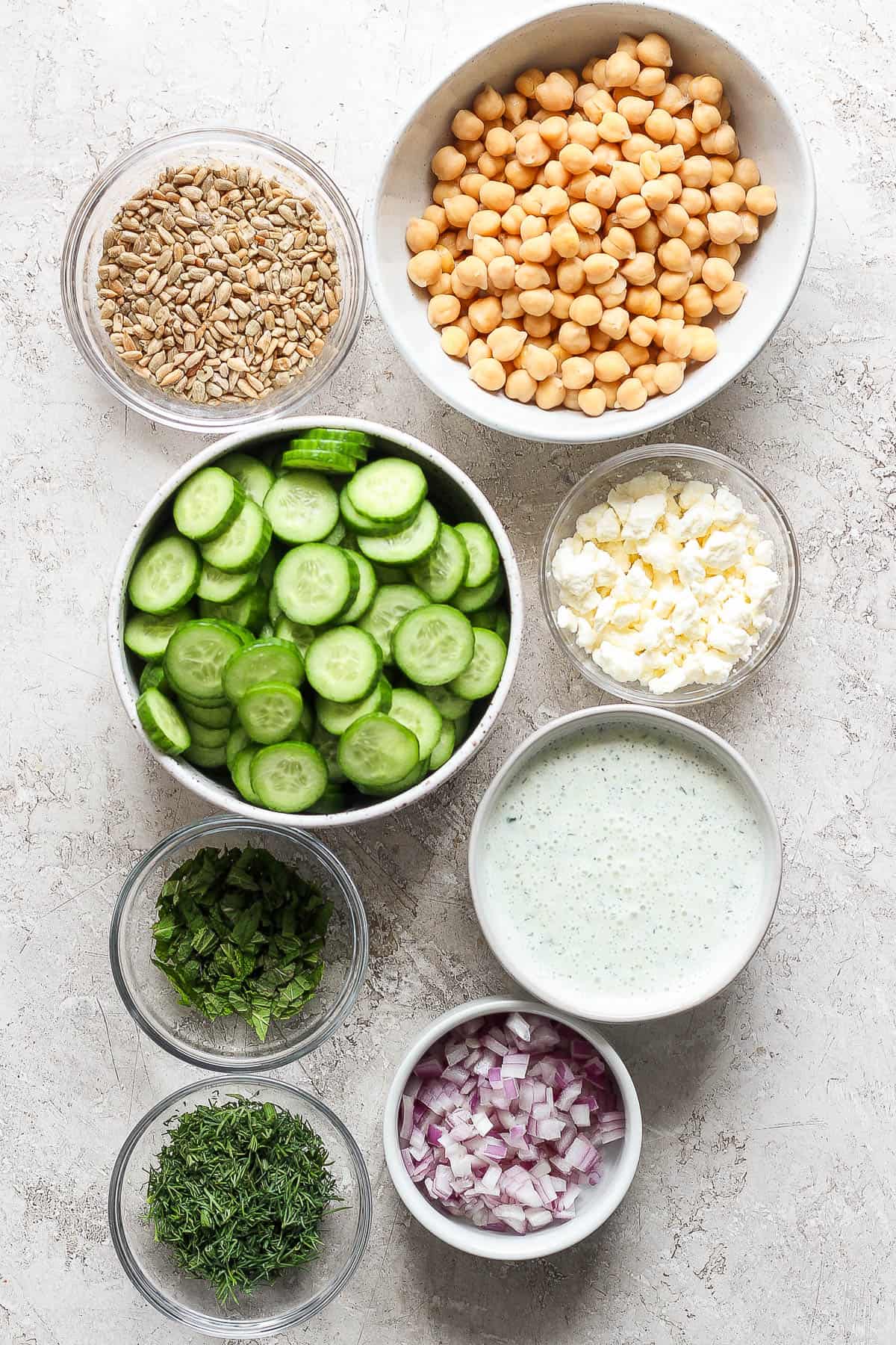 Individual bowls of chickpeas, sunflower seeds, sliced cucumbers, feta, chopped mint, fresh dill, diced red onion, and tzatziki dressing.