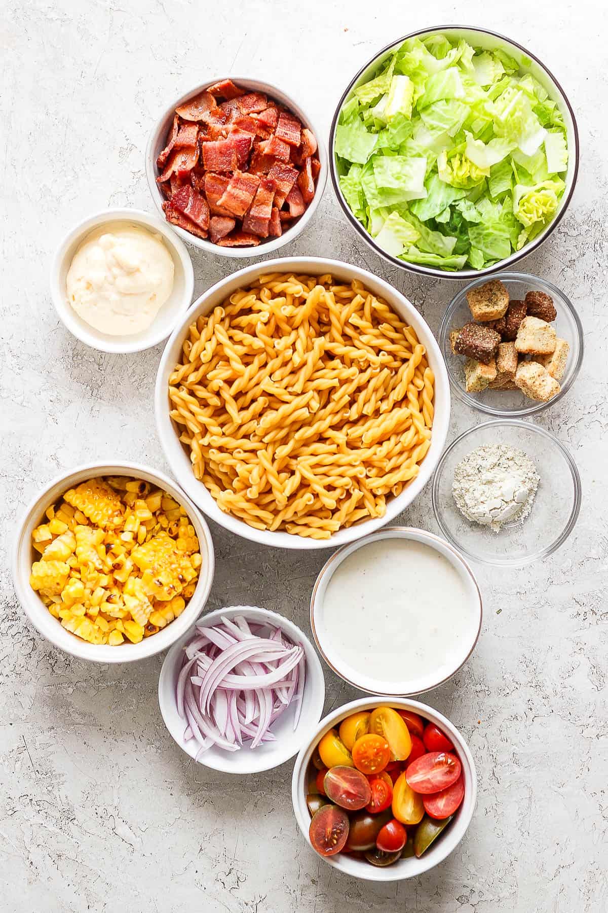 Individual bowls of chopped romaine, chopped bacon, croutons, ranch seasoning, grilled corn, sliced red onions, halved cherry tomatoes, gemeli noodles, ranch dressing, and mayo. 