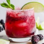 A glass filled with a blackberry mojito with blackberries next to it and garnished with fresh mint leave and lime wedge.