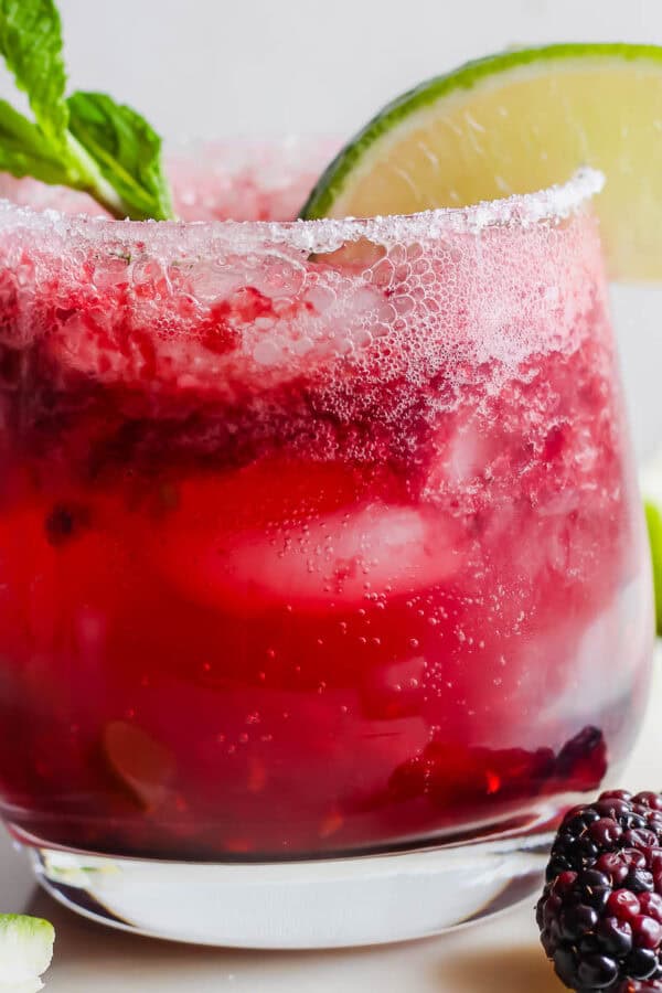 A glass filled with a blackberry mojito with blackberries next to it and garnished with fresh mint leave and lime wedge.