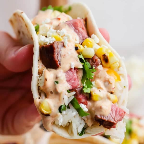 Someone holding a grilled steak taco with cilantro lime rice, grilled corn, steak and chipotle lime sauce.