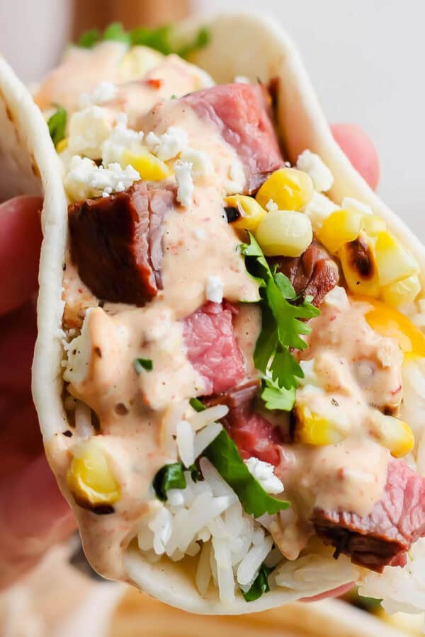 Someone holding a grilled steak taco with cilantro lime rice, grilled corn, steak and chipotle lime sauce.