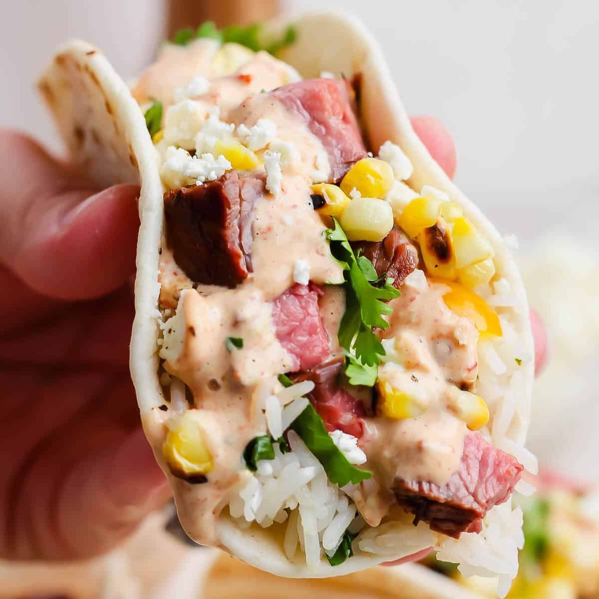 Someone holding a grilled steak taco with cilantro lime rice, grilled corn, steak and chipotle lime sauce.