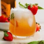 A glass of a Kentucky Buck with ice inside and a strawberry next to it and sitting on the rim with a bottle of ginger beer in the background.