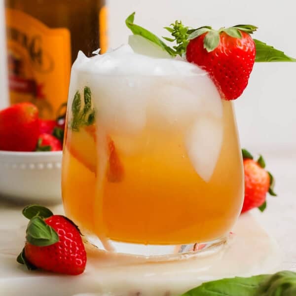 A glass of a Kentucky Buck with ice inside and a strawberry next to it and sitting on the rim with a bottle of ginger beer in the background.