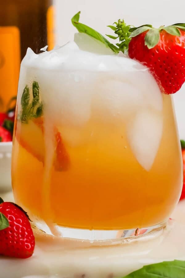 A glass of a Kentucky Buck with ice inside and a strawberry next to it and sitting on the rim with a bottle of ginger beer in the background.