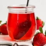 A small jar of strawberry basil simple syrup with a spoon sticking out and strawberries around it.