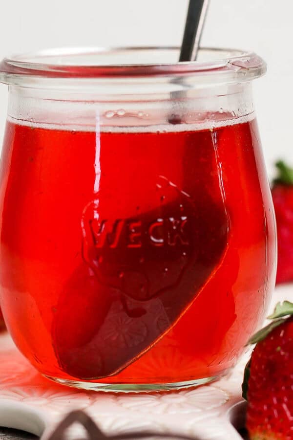A small jar of strawberry basil simple syrup with a spoon sticking out and strawberries around it.