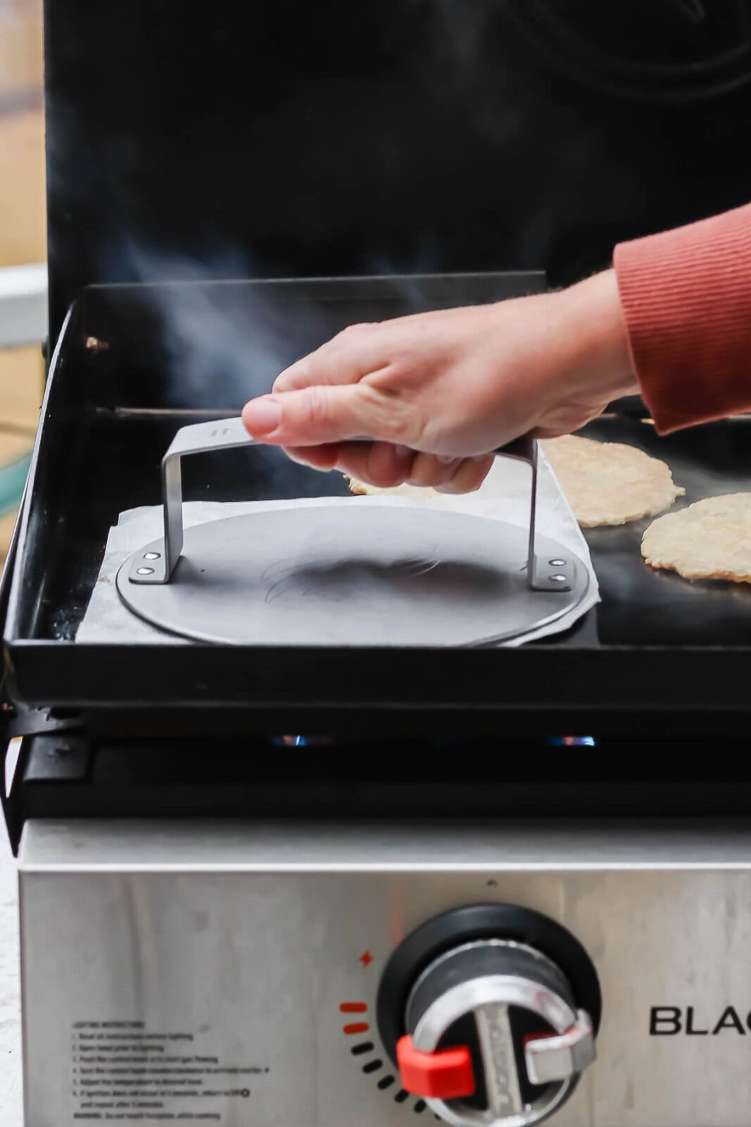A hand pressing a burger press flattening a chicken patty on a cooktop.