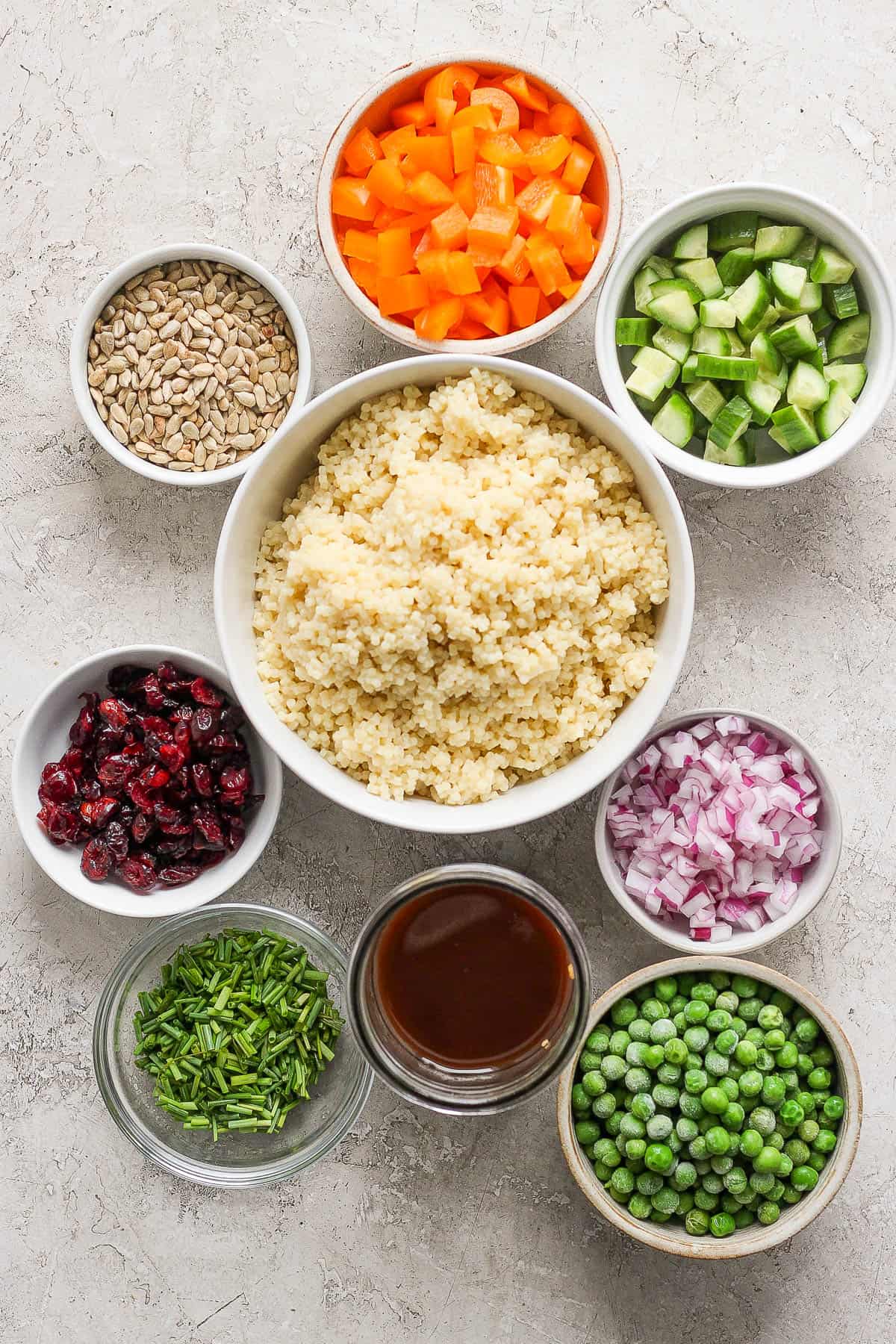 Individual bowls of chopped bell pepper, chopped cucumber, cooked acini di pepe, sunflower seeds, cranberries, diced red onion, chopped chives, frozen peas, and a salad dressing.