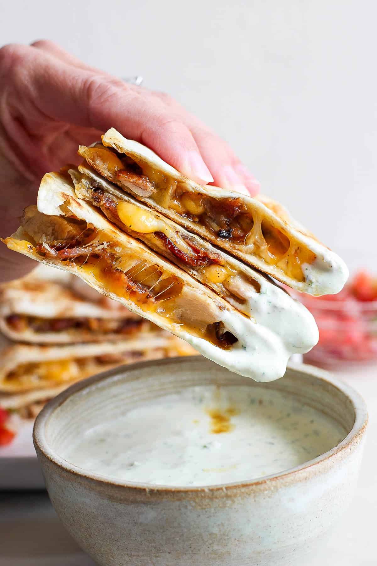 A hand holding three chicken bacon ranch quesadillas with a scoop of jalapeno ranch on one end.