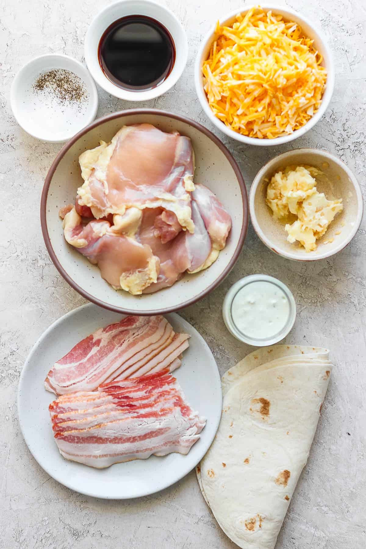 Individual bowls of soy sauce, salt and pepper, shredded cheese, chicken thighs, garlic butter, jalapeno ranch, bacon, and tortillas.