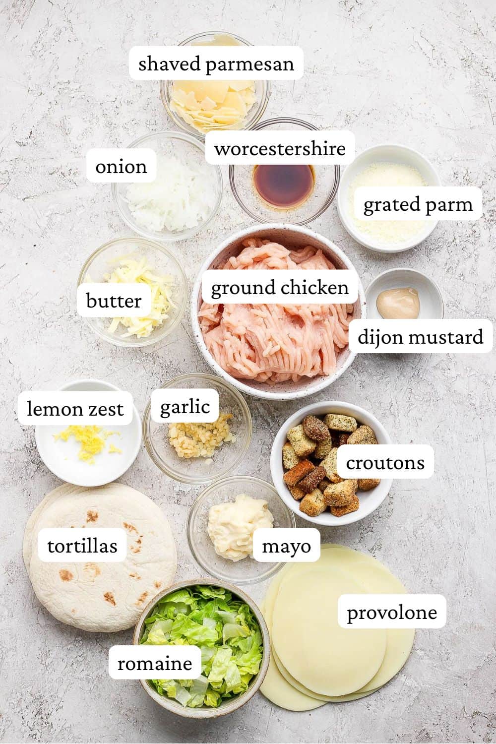 Individual bowls of shaved parmesan cheese, shaved onion, grated parmesan cheese, grated butter, ground chicken, dijon mustard, minced garlic, lemon zest, croutons, mayo, chopped romaine, tortillas, white cheese, and worcestershire.