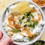 Someone holding a bowl of fried pickle dip with chopped pickles on top and potato chips sticking out.