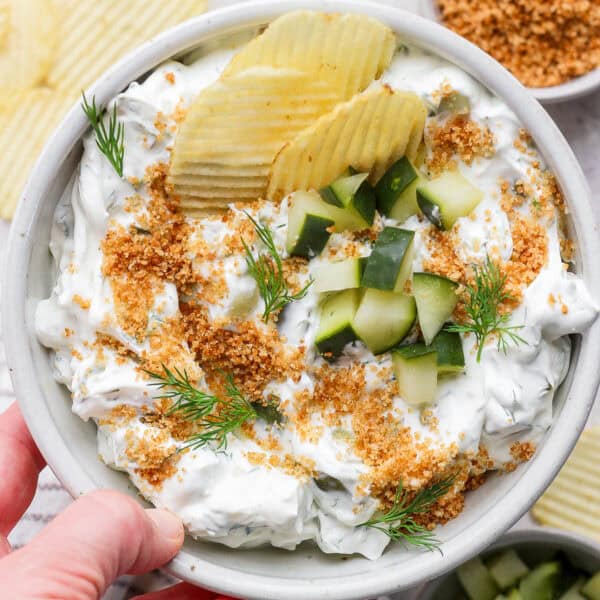 Someone holding a bowl of fried pickle dip with chopped pickles on top and potato chips sticking out.