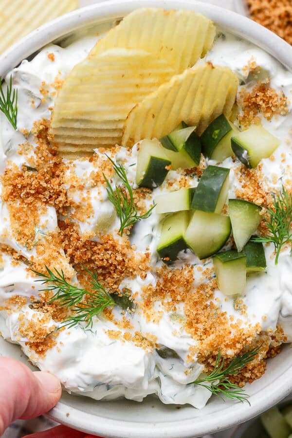 Someone holding a bowl of fried pickle dip with chopped pickles on top and potato chips sticking out.