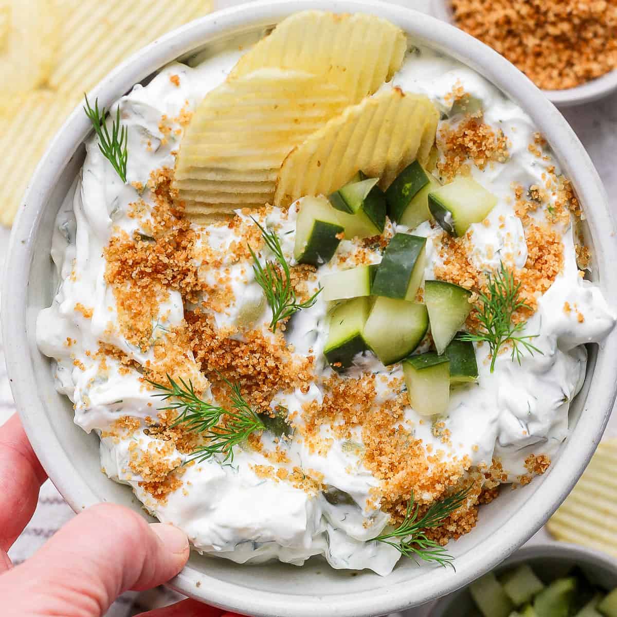 Someone holding a bowl of fried pickle dip with chopped pickles on top and potato chips sticking out.