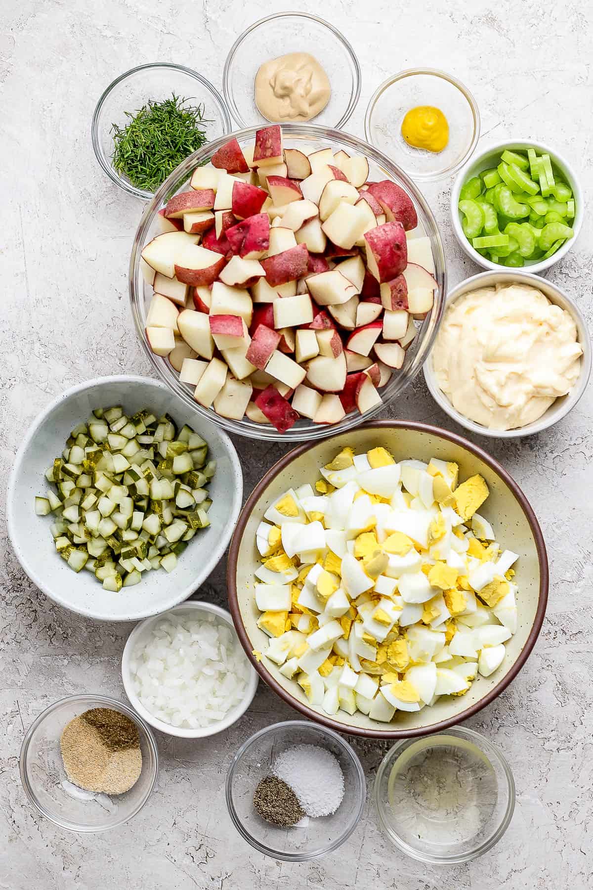 Individual bowls of red potatoes, dijon mustard, yellow mustard, sliced celery, chopped fresh dill, diced pickles, mayo, chopped hard boiled eggs, diced yellow onion, salt and pepper, vinegar, and garlic powder, onion powder, and celery salt.