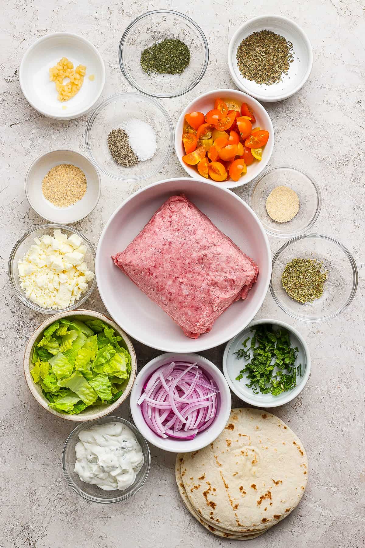 Individual bowls of salt and pepper, minced garlic, quartered cherry tomatoes, onion powder, garlic powder, feta cheese, ground lamb, chopped romaine lettuce, sliced red onion, chopped fresh mint, dried oregano, dried basil, dried dill, homemade tzatziki sauce, flour street taco tortillas.