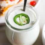 A jar of homemade jalapeño ranch with spoon sticking out and a slice of jalapeño on top.