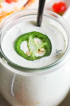 A jar of homemade jalapeño ranch with spoon sticking out and a slice of jalapeño on top.