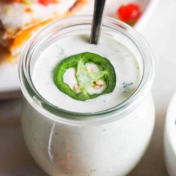 A jar of homemade jalapeño ranch with spoon sticking out and a slice of jalapeño on top.