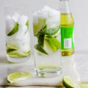 A glass of rose's lime juice gin and tonic with limes along with a bottle of rose's lime juice.