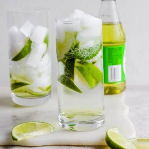 A glass of rose's lime juice gin and tonic with limes along with a bottle of rose's lime juice.