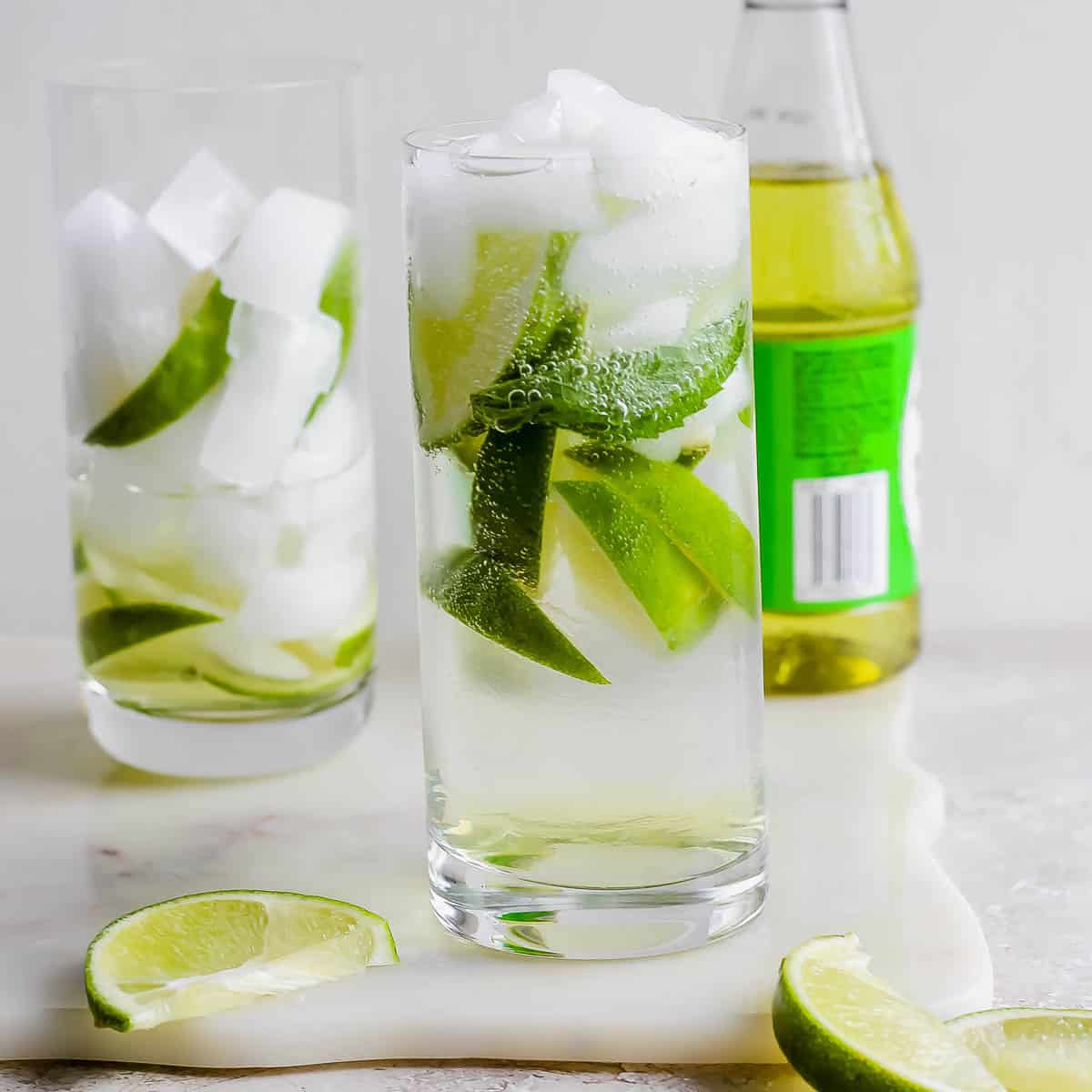 A glass of rose's lime juice gin and tonic with ice and limes inside with another in the background along with a bottle of rose's lime juice.