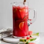 A pitcher of watermelon margaritas with ice, limes and watermelon slices inside and a spoon sticking out.