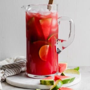 A pitcher of watermelon margaritas with ice, limes and watermelon slices inside and a spoon sticking out.