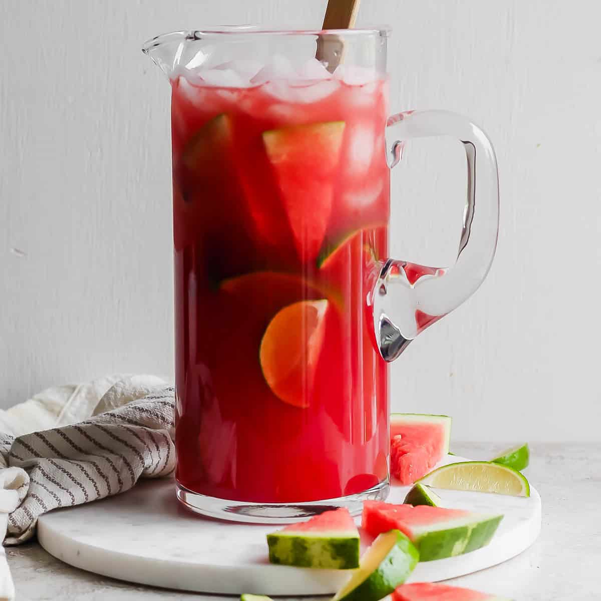 A pitcher of watermelon margaritas with ice, limes and watermelon slices inside and a spoon sticking out.