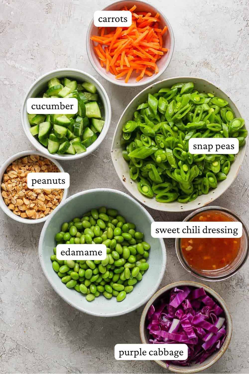 Sugar snap pea salad + sweet chili vinaigrette ingredients in individual bowls including matchstick carrots, cucumber, snap peas, peanuts, edamame, sweet chili dressing, and purple cabbage.