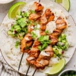 A plate of rice with three grilled bang bang chicken skewers on top, bang bang sauce and avocado cucumber salad on a plate with lime wedges.
