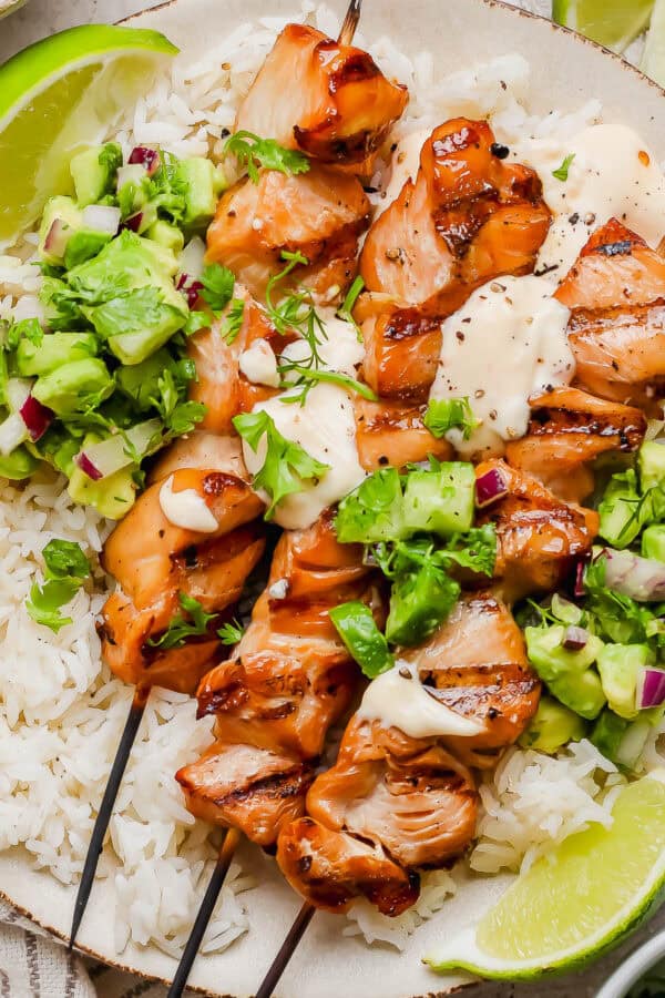 A plate of rice with three grilled bang bang chicken skewers on top, bang bang sauce and avocado cucumber salad on a plate with lime wedges.