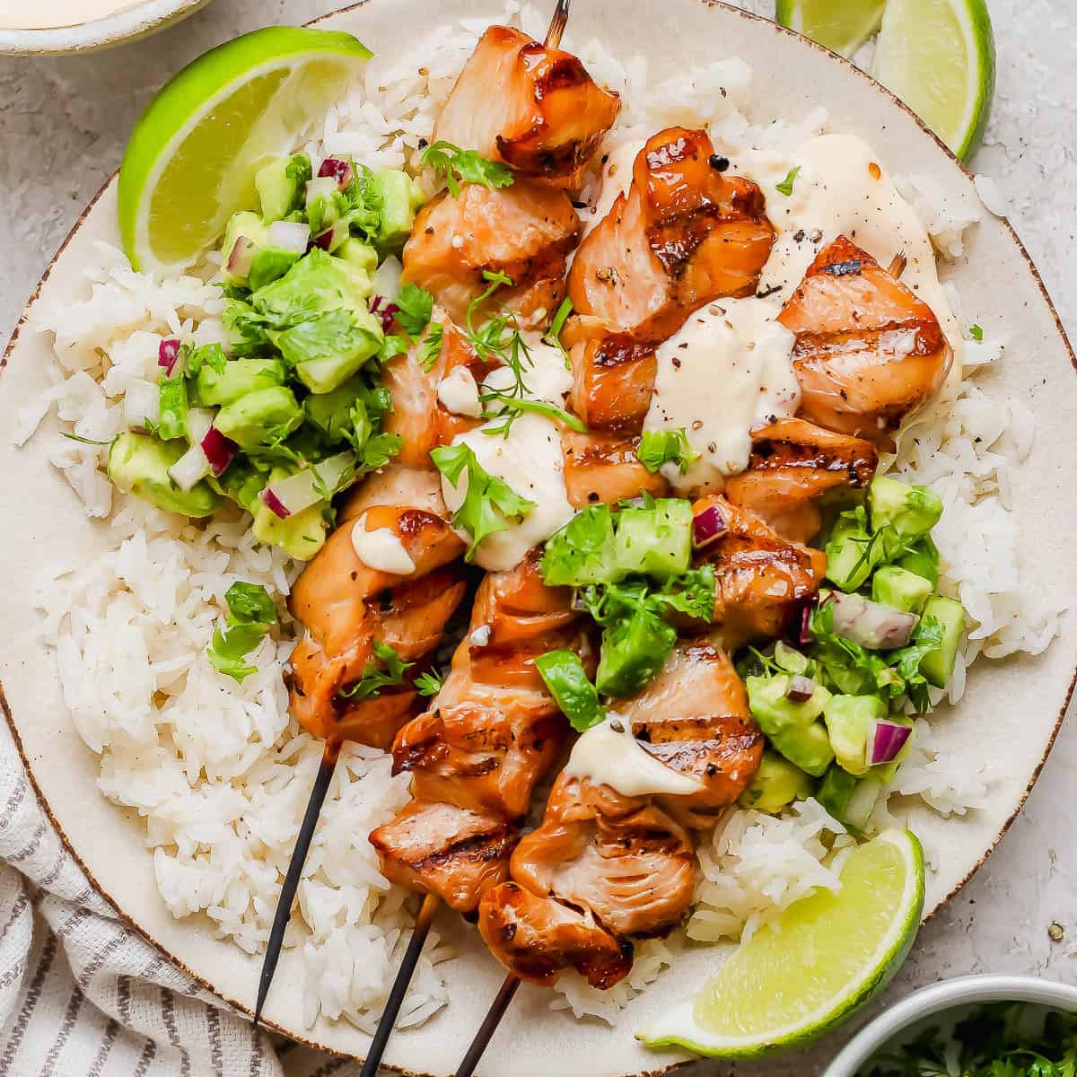 A plate of rice with three grilled bang bang chicken skewers on top, bang bang sauce and avocado cucumber salad on a plate with lime wedges.