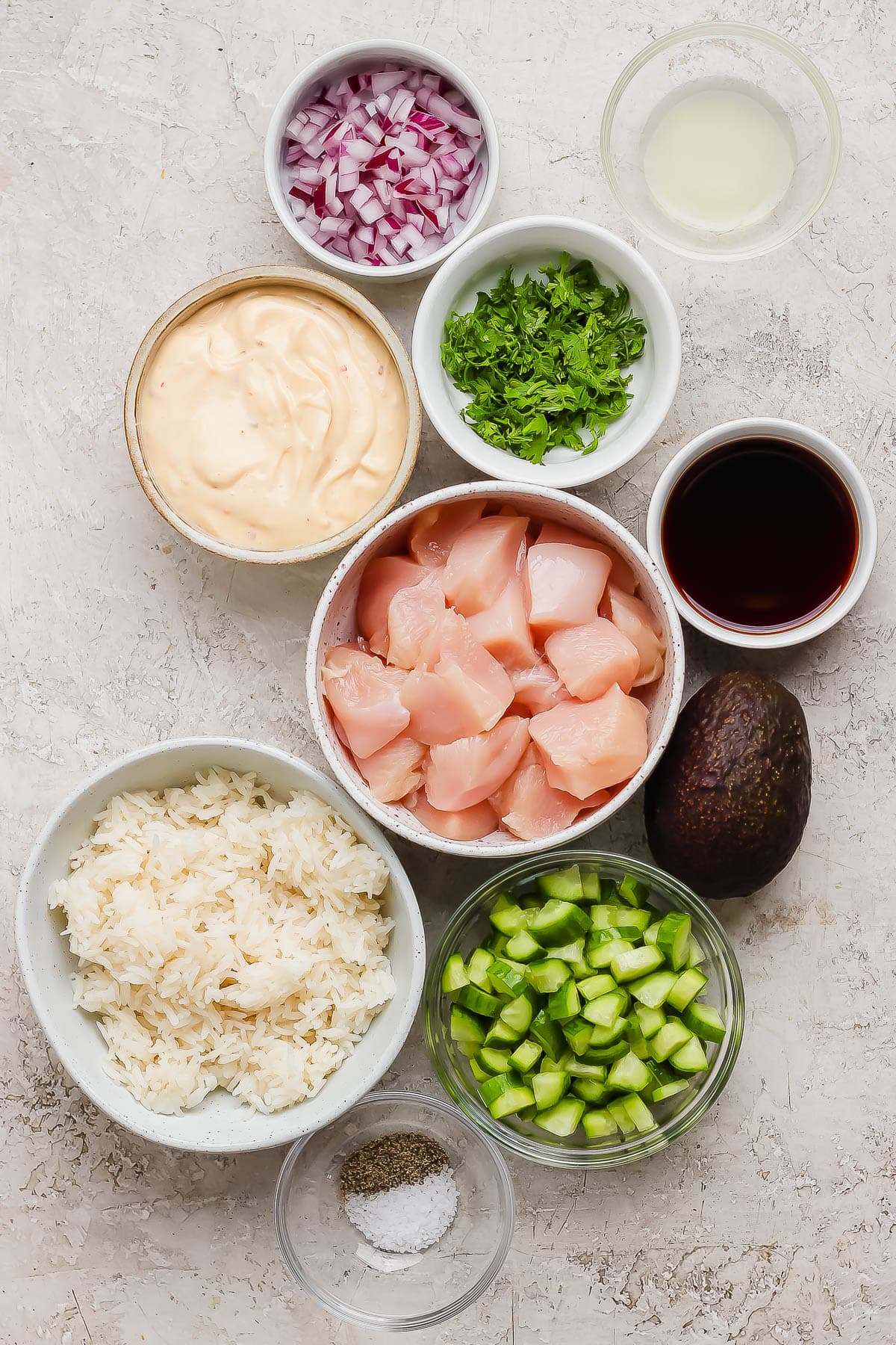 Individual bowls of lime juice, diced red onion, cilantro. bang bang sauce, soy sauce, cubed chicken breasts, cooked white rice, sliced and quartered cucumbers, salt & pepper, and a whole avocado.