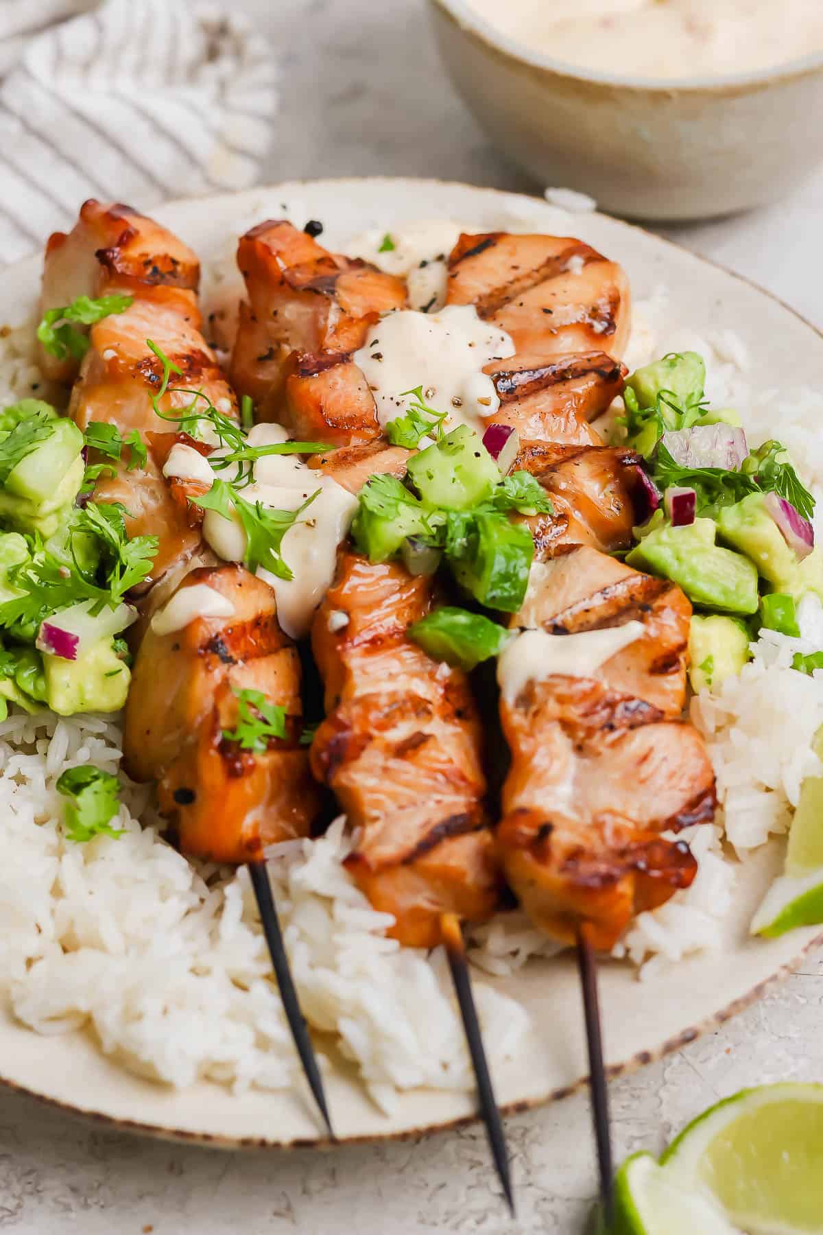 Three chicken skewers on a plate on top of rice topped with avocado salsa and bang bang sauce.