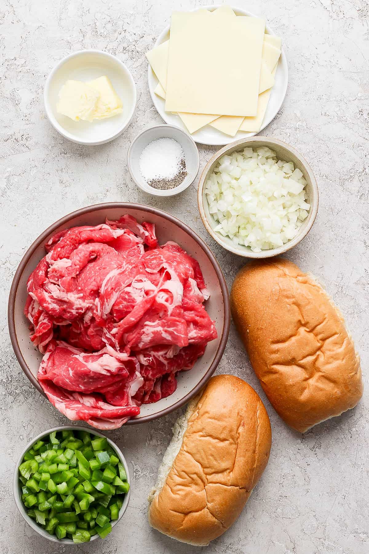 Individual bowls of butter, salt and pepper, diced onion, slices of white American cheese, thinly sliced ribeye steak, diced green peppers, and two hoagie rolls.